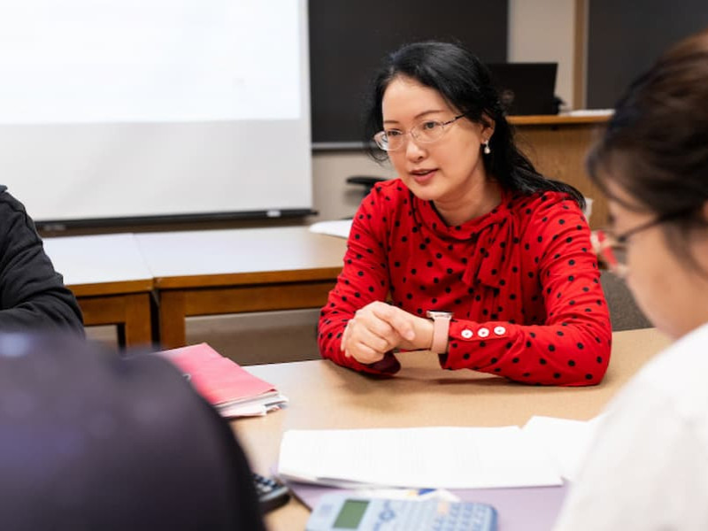 A female instructor wearing glasses and a red polka-dot blouse speaks with students seated around a table in a classroom setting.