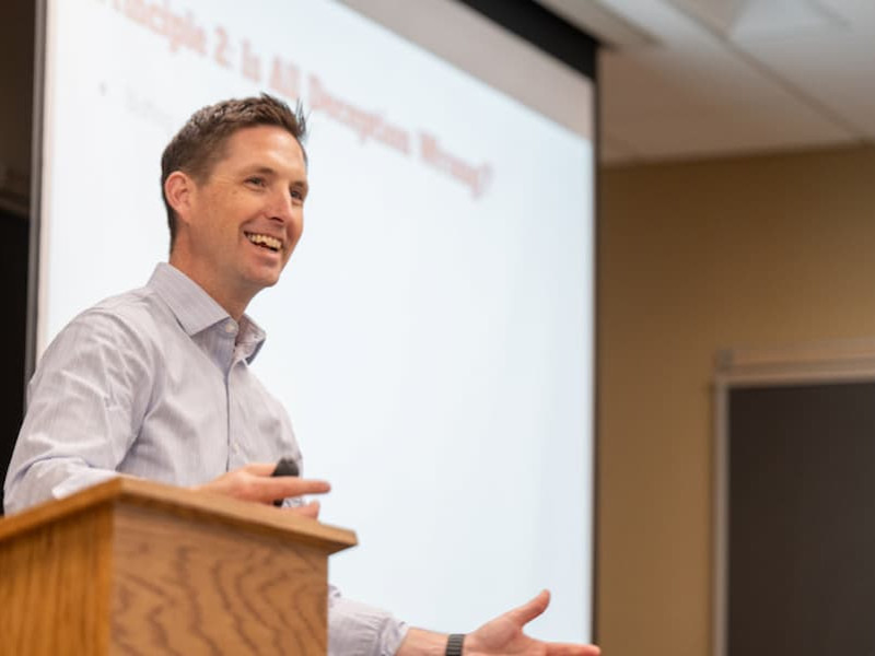 A smiling male instructor speaks behind a wooden podium in a classroom, with a presentation slide projected on the screen behind him.