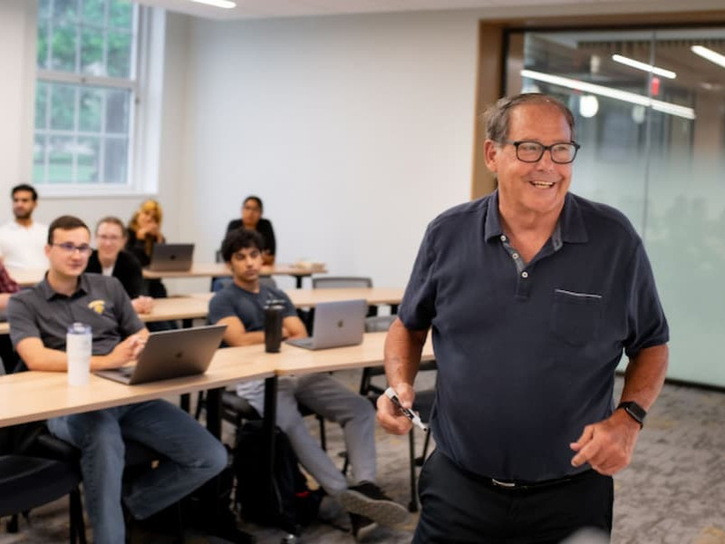 A smiling professor holding a dry-erase marker and speaking in front of a class of university students seated at desks with laptops.