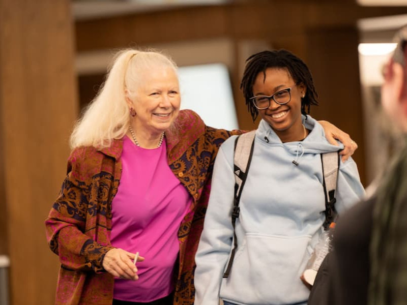 A smiling female professor with her arm around a student's shoulder as they talk indoors on campus.