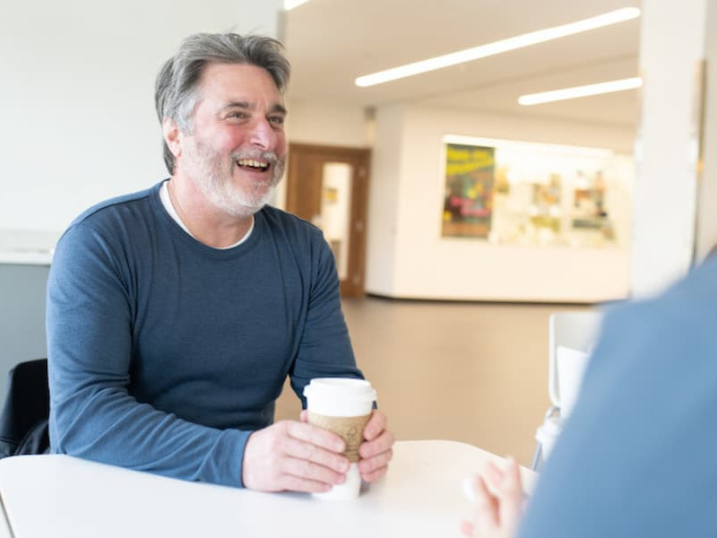 A smiling, bearded man holding a coffee cup while sitting and talking with someone at an indoor table.