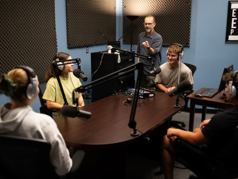 Students wearing headphones sit around a table with microphones in a recording studio while an instructor speaks to them during a podcasting class.