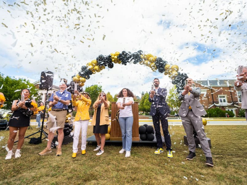 A group of people smiling and posing together behind a podium at an outdoor podium ceremony on a university campus.