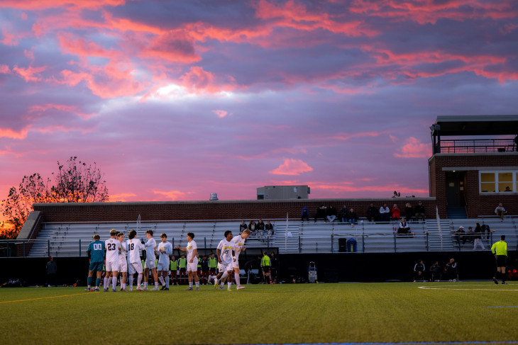 DePauw University soccer players huddle on Cotton Field with Reavis Stadium and a pink sunset in the background.