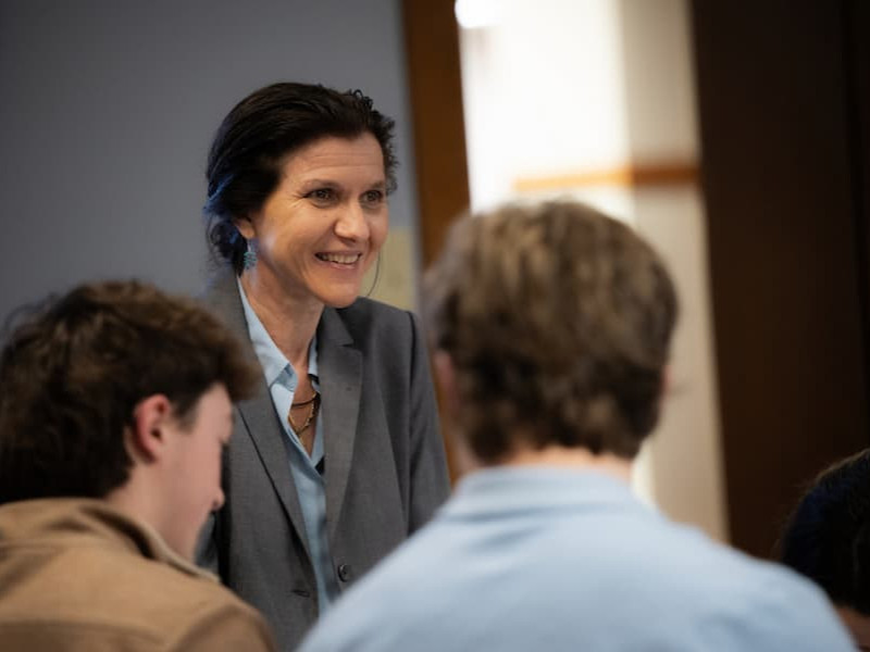 A female professor smiling and gesturing with her hands while speaking to a group of students in a brightly lit campus lounge or library.