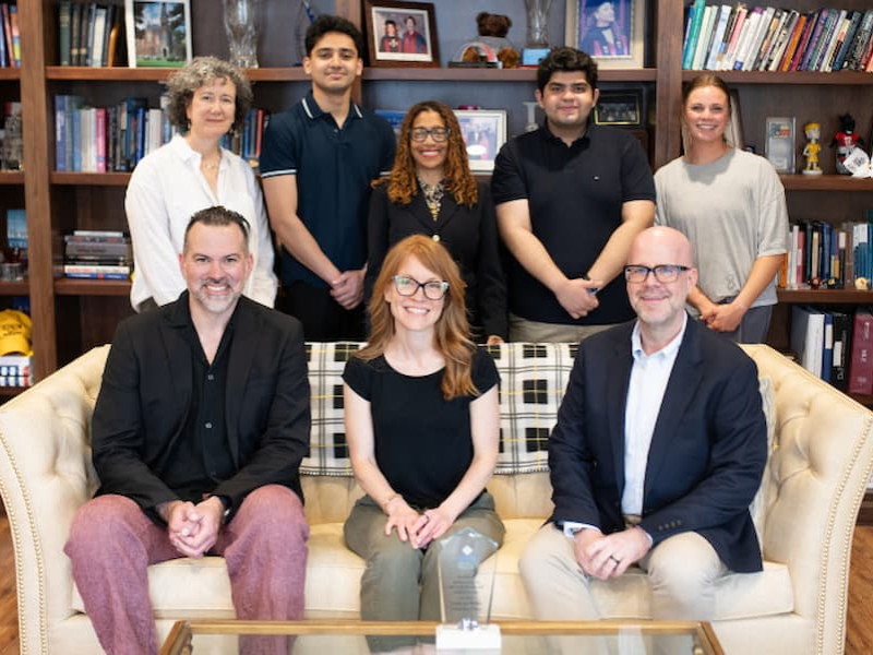 A diverse group of students standing shoulder to shoulder and smiling warmly in a brightly lit indoor lounge or campus center.