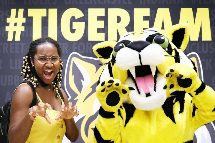 DePauw student posing with the DePauw Mascot, Tyler the Tiger. The two are in front of a black and gold DePauw Tiger Family banner on-campus.