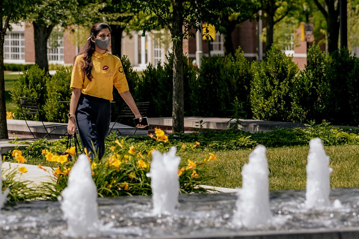 Woman walks on campus behind fountain and flowers