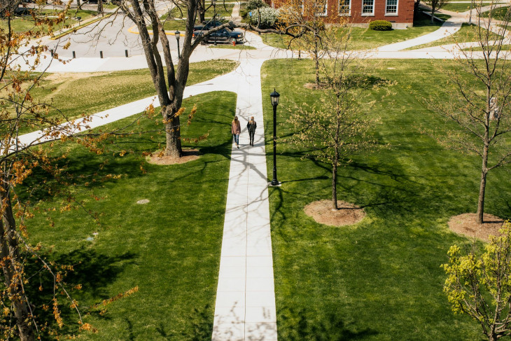 Two students walk along a sidewalk amidst budding trees on campus