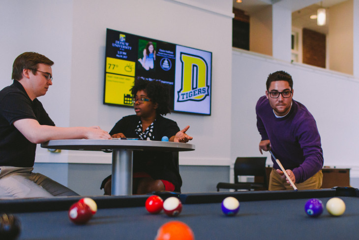 faculty and student talking at a table in front of a digital sign. student in foreground playing pool. 