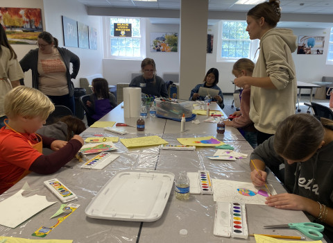 A group of DePauw students and elementary school children are sitting around a table working on abstract watercolor paintings.