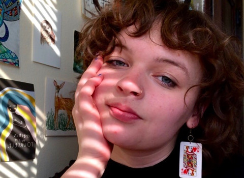 Nina faces the camera next to a wall of artwork with light shining through blinds on her face and the wall.