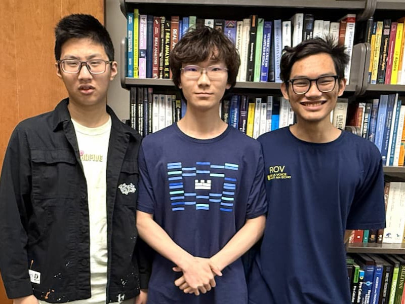 Three male students wearing glasses stand side by side in front of bookshelves filled with textbooks.