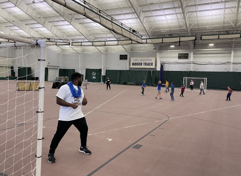 A student is standing by a soccer goal post in an indoor track. He is smiling and looking back at some young children behind him. They appear to be playing a running game and having lots of fun.