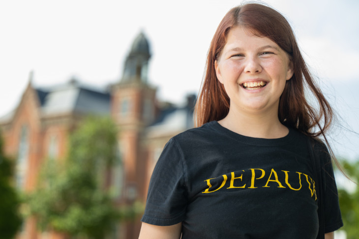 View of campus with a DePauw University student smiling towards you. Historic East College on DePauw's campus sits in the background.