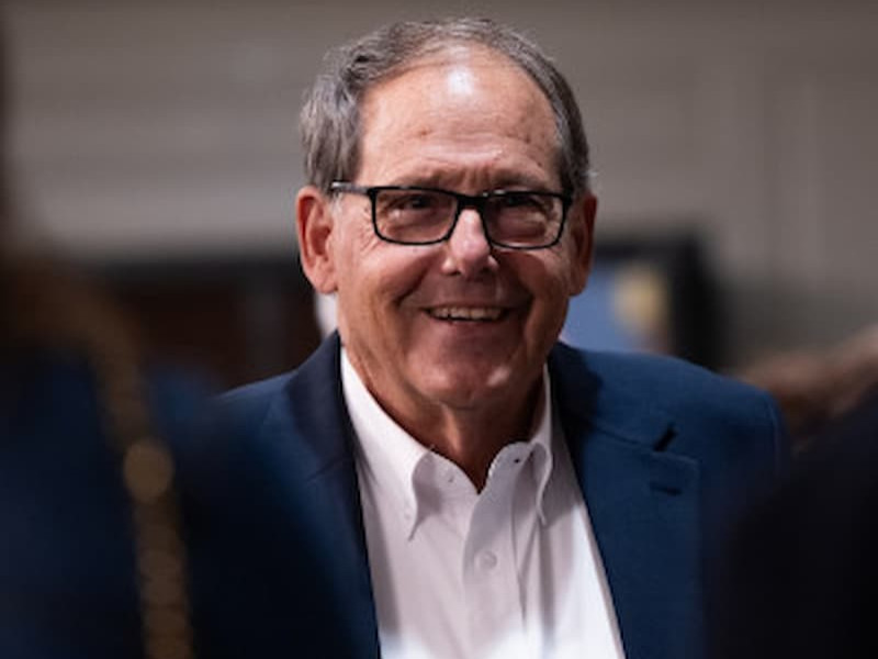 A smiling man wearing glasses, a white dress shirt, and a blue blazer looking forward at an indoor event.