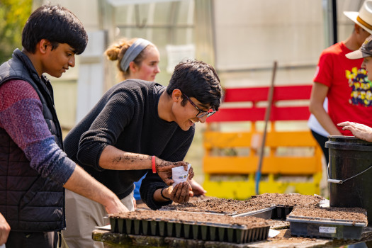 A student smiles as he plants seeds into a plastic seed tray. His hands are covered in soil and his classmates fill the background.