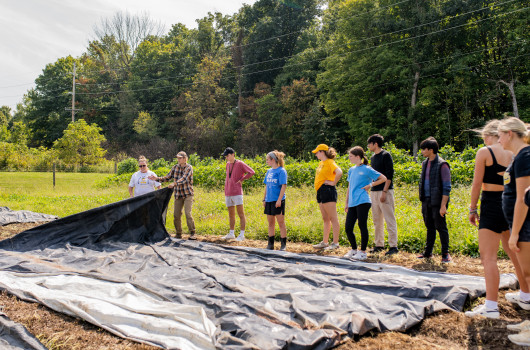 A group of students stand around a black silage tarp in the farm field on bare soil, learning how to move the tarp.