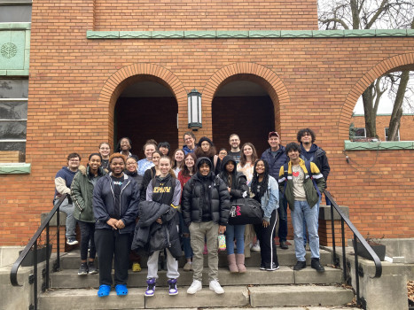 A group of students in winter clothes are standing on the steps of a brick building smiling at the camera.