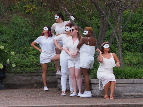 A group of feminine people dressed in white with white eye masks look off to their left