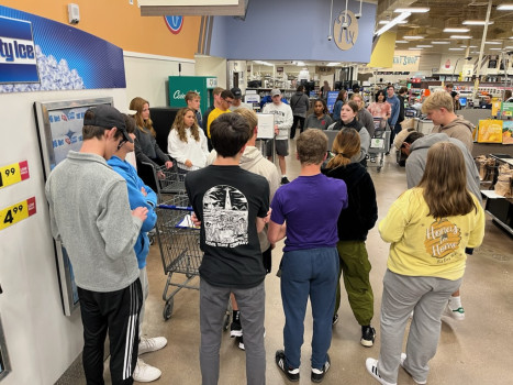 Students are gathered around our service coordinator in the entrance of a Kroger. She is wearing a black sweater and black scarf and explaining the rules of Supermaket Sweep.
