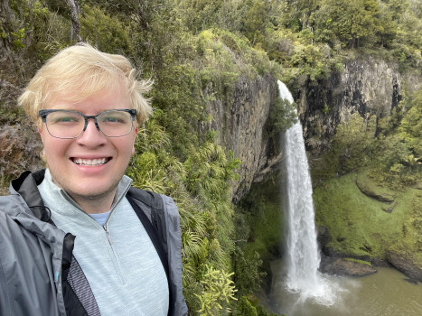A male student with blond hair and glasses is smiling in front of a waterfall in a green forest.