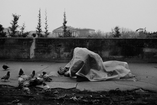 Pigeons surrounding a person sitting on the sidewalk covered by a plastic tarp.