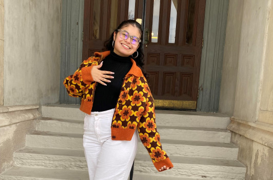 A female student is standing on the stone steps of East College. She is wearing a brown retro floral jacket and smiling at the camera.