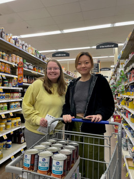 Two female students are standing behind a shopping cart at Kroger, full of supplies they have picked up for Supermarket Sweep.