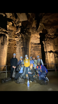 A group of students are standing in a dark cave, posing for a group photo. Some of them have on hard hats with lamps.