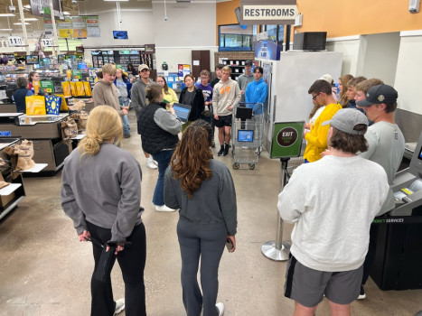 A group of students are standing in a circle in front of an ice machine in the Kroger entranceway. They are listening to instructions for Supermarket Sweep 2023.
