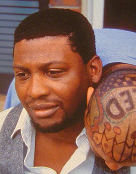 Seated man wearing a white shirt and grey vest while another man is leaning over his shoulder showing tattoos on his bald head