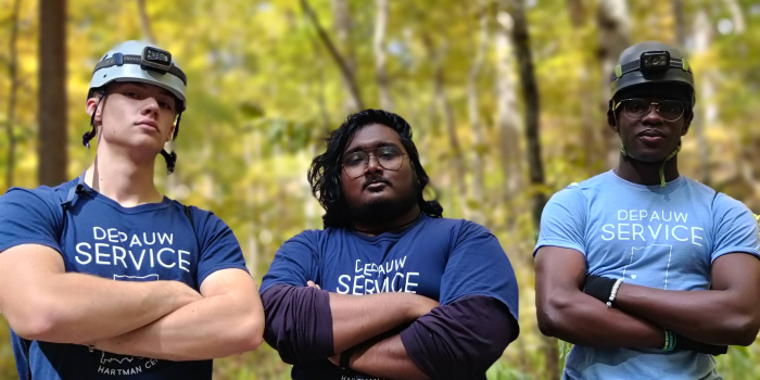 Three male students are standing with their arms crossed over their chests. They are standing in the woods. All three of them are wearing blue DePauw Service shirts and looking proud.