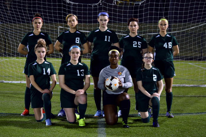 A group of women pose seriously on a soccer field wearing green uniforms.