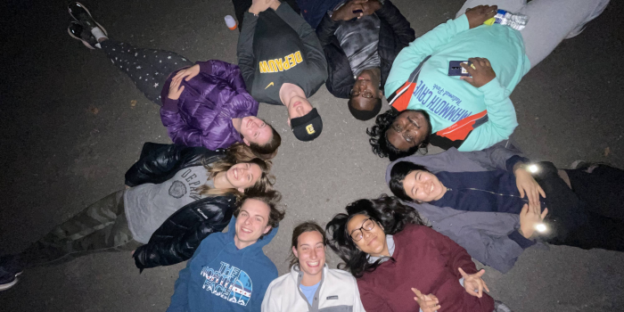 A group of university students lying in a circle with their heads pointed towards the center. They are lying down on the ground in the dark, stargazing. Everyone is smiling.