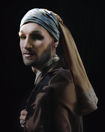 A man with a beard wearing a long pearl earring, a blue turban wrapped around his head with a yellow piece of fabric coming out of it in the back that has lace around the bottom edge
