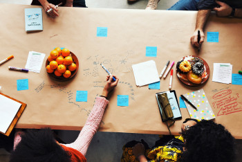 People sitting at a table that has plates with oranges and donuts will writing on a table covered with brownish paper