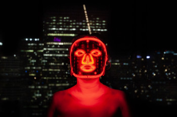 A person in a electronic glowing mask and red lights in front of a night time cityscape