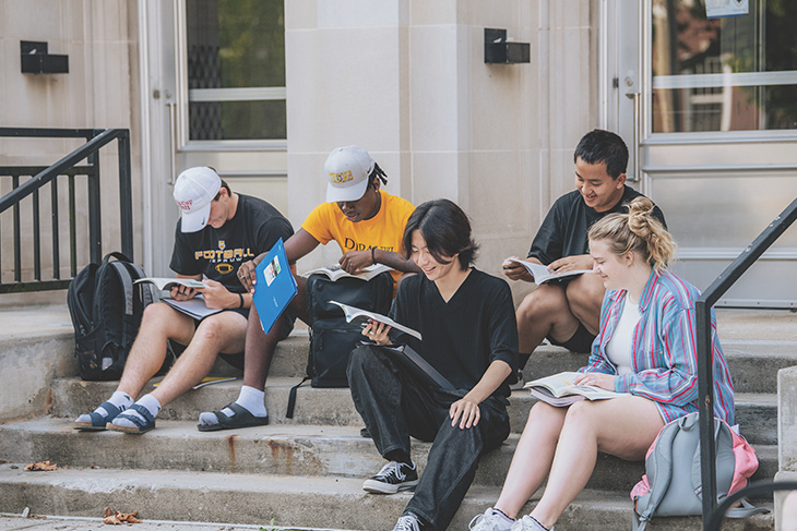 Four students seated on the stairs in front of Harrison hall, during an outdoor class lecture