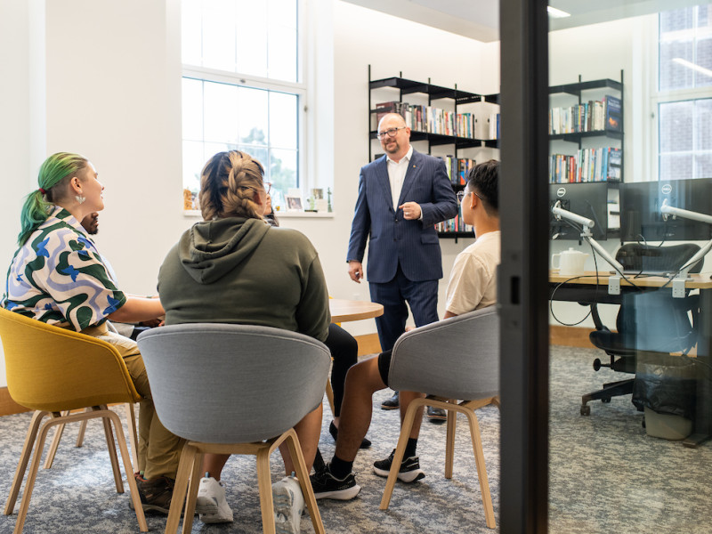 Man speaking with three students in a bright campus study room