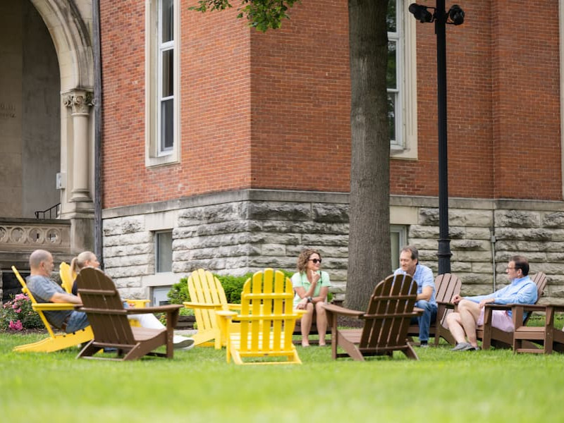 A group of alumni sit in a circle of yellow and brown Adirondack chairs on a green lawn near a brick building.