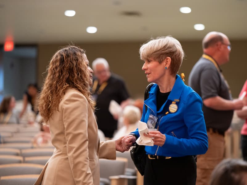 Two women stand and talk closely in an auditorium while other event attendees mingle in the background.