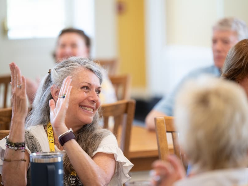 A woman with grey hair laughs and raises her hand while seated at a wooden table during an indoor event.