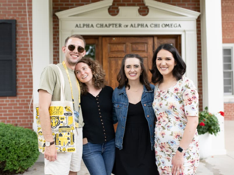 Four young alumni smiling together in front of the Alpha Chapter of Alpha Chi Omega entrance.
