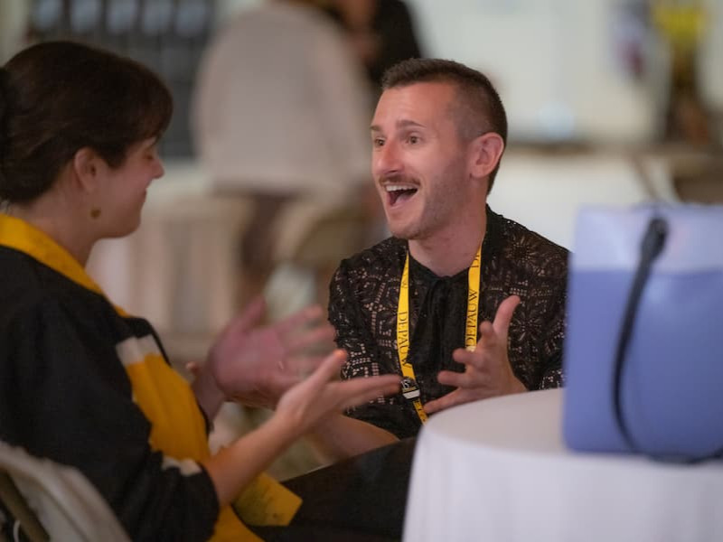 A man wearing a yellow lanyard expresses excitement during a conversation at a table with another guest.