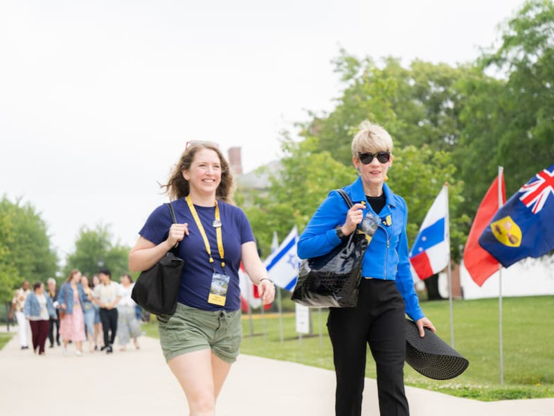 Two women walk along a campus path lined with international flags, wearing yellow event lanyards.