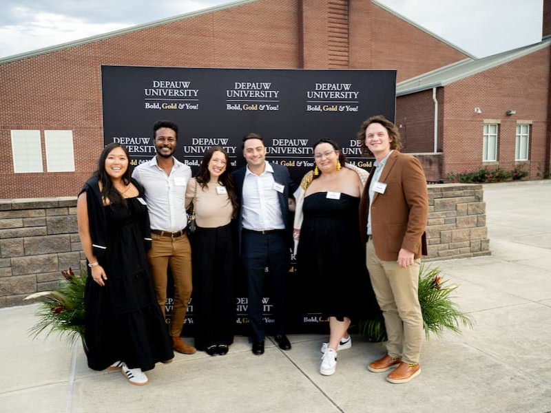 Six smiling alumni posing together in front of a DePauw University Bold, Gold & You! backdrop.