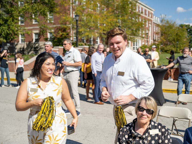 Two people holding yellow and black pom-poms smile while walking through an outdoor event crowd on campus.