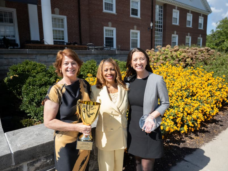 Three women posing outdoors while holding a large gold trophy and a glass award.