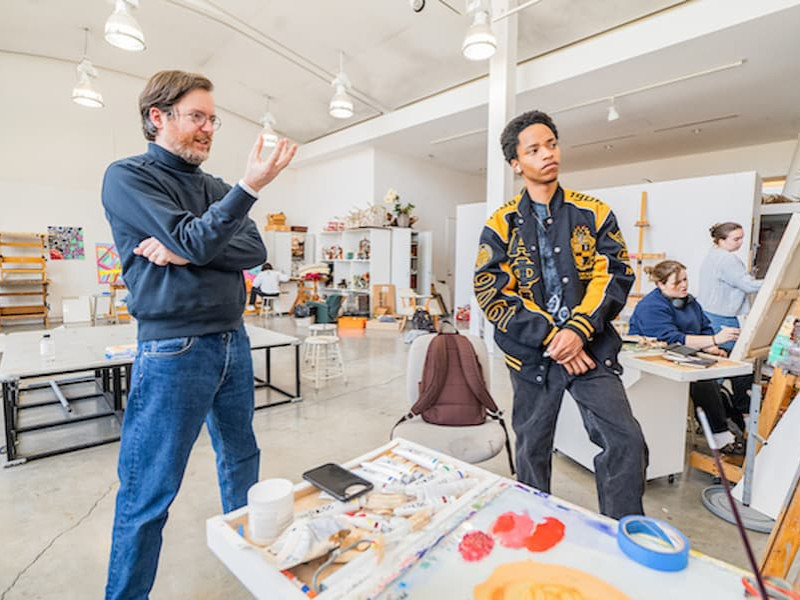 An art instructor talking to a student in a bright studio filled with easels and painting supplies.